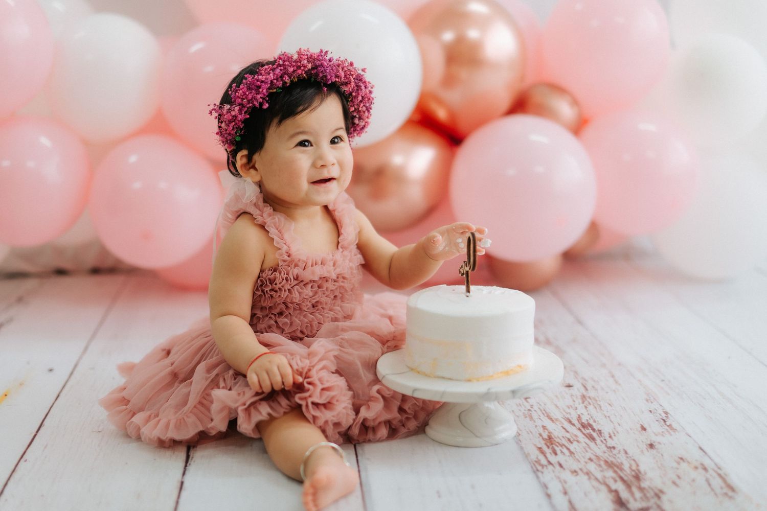 Baby in pink tulle dress and floral crown sits on wooden floor with white cake and pink balloon backdrop.