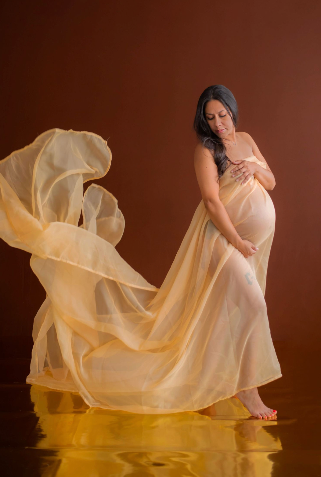 Maternity photo of woman in flowing champagne dress against brown backdrop.