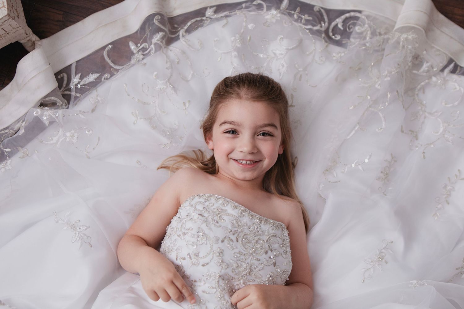 A young child in a white lace dress poses elegantly on a soft white backdrop during a portrait session.