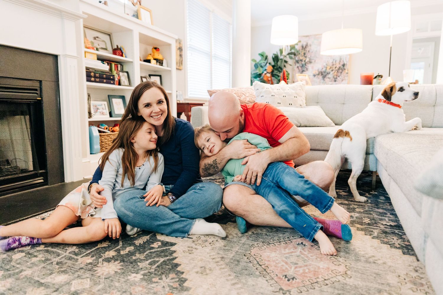 A playful photo of a Charlotte family and a white dog laying on a gray carpet in a bright living room.