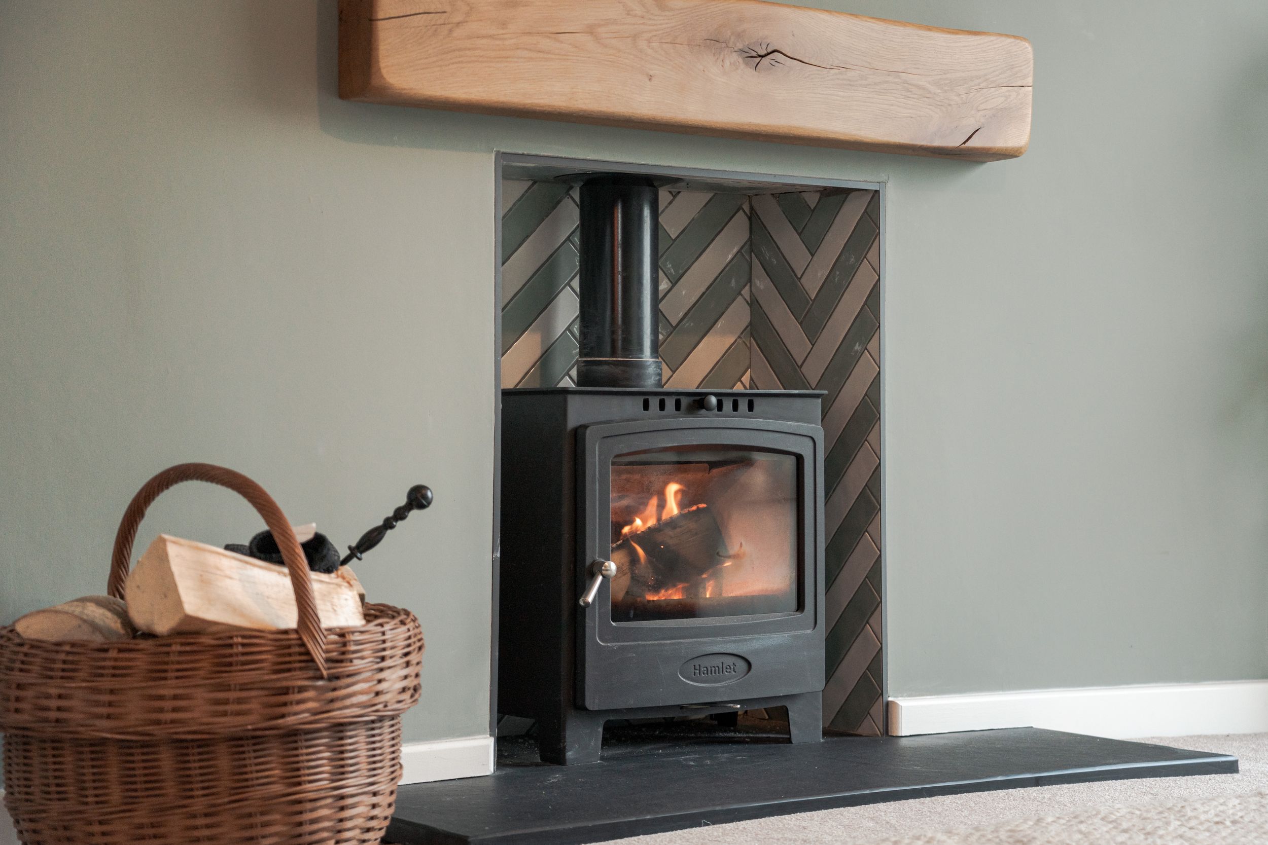 Rustic fireplace setup with woven basket and wooden mantel against herringbone pattern tile backdrop.