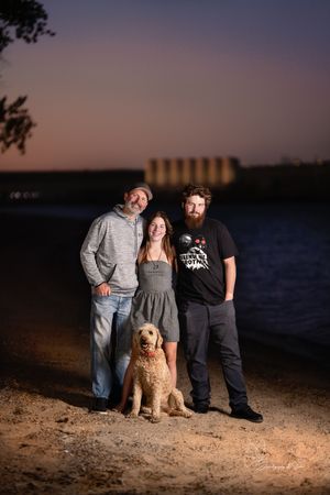 A group poses with their goldendoodle dog at sunset near a lake with grain silos in the background in Pierre, South Dakota.