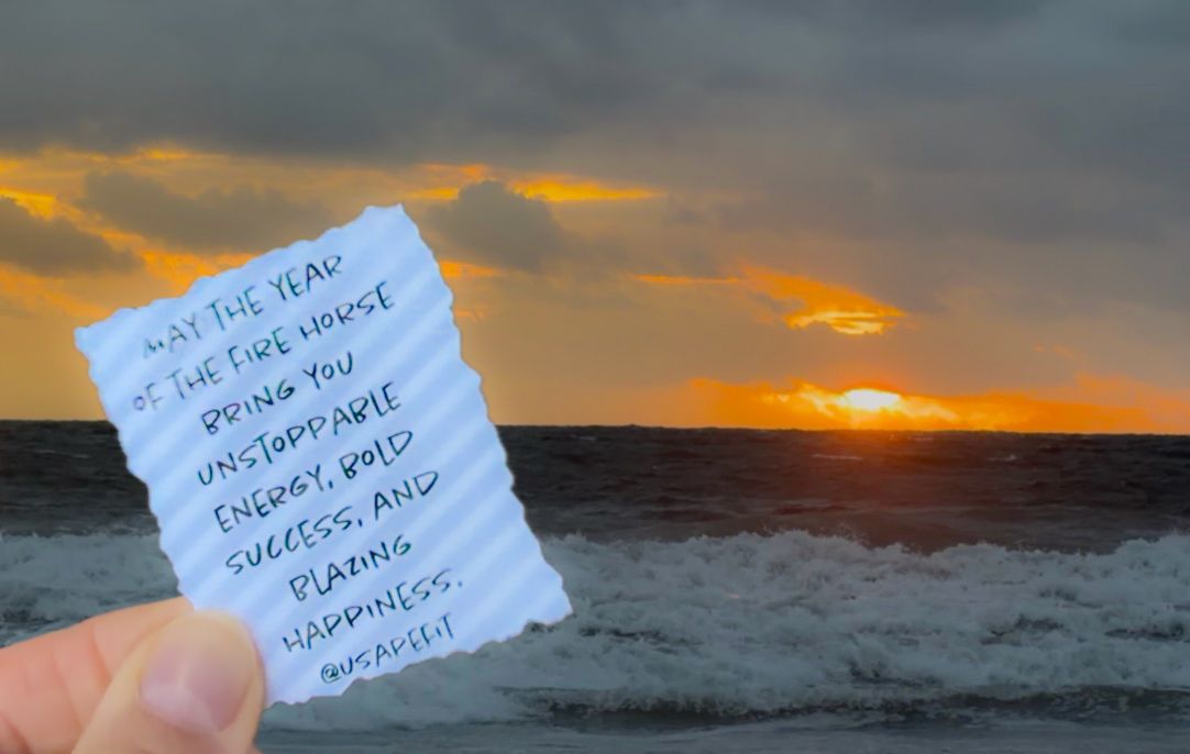 A handwritten note held against a vibrant orange sunset over the ocean horizon.