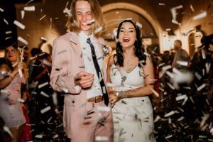 Wedding celebration with guests dancing and confetti flying in warm-lit indoor reception venue.