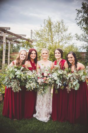 Bridal party in burgundy dresses and white gowns pose with lush greenery bouquets outdoors.