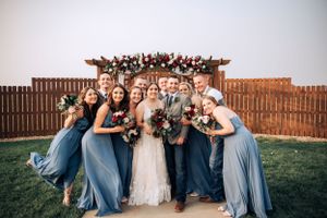 Wedding party in blue dresses and suits poses outdoors with white fence backdrop.
