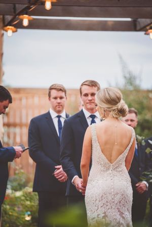 A wedding ceremony setup with string lights overhead and guests in formal attire gathered outdoors.