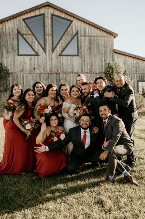 Large wedding party in burgundy dresses and dark suits posing together by rustic barn.