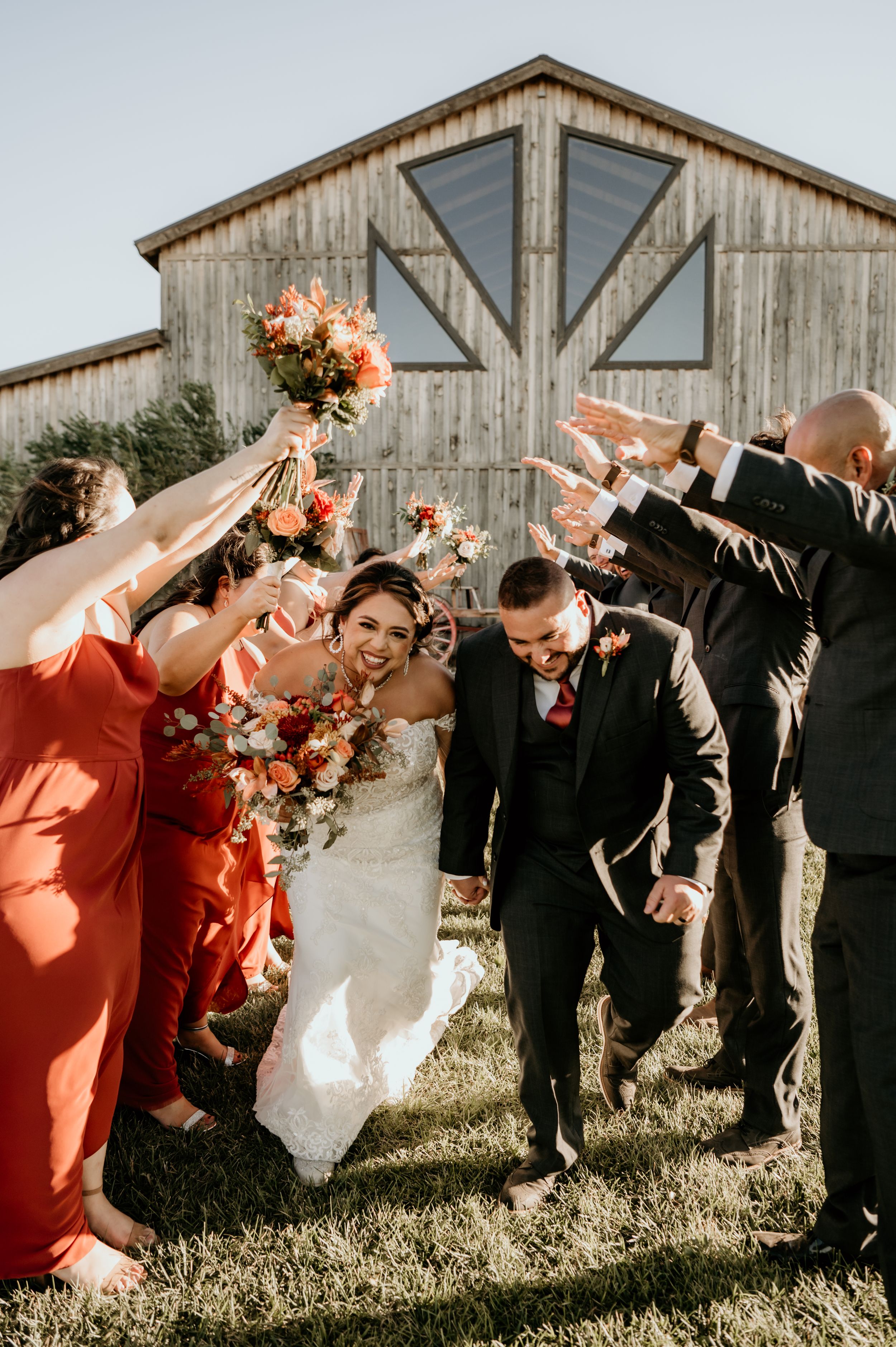 Wedding party in coral dresses and black suits pose playfully in front of rustic barn.