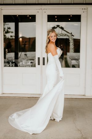 Elegant bride in modern white wedding dress twirls playfully outside a white building.