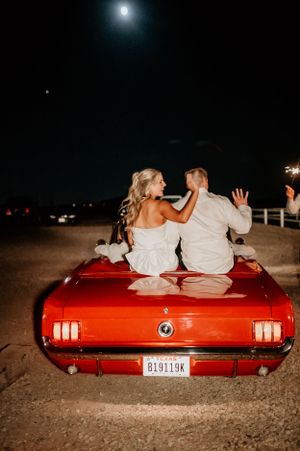Couple sits on red vintage car under moonlight for romantic wedding photo.