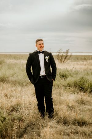 Groom in classic black tuxedo poses in rustic outdoor field during golden hour wedding photoshoot.