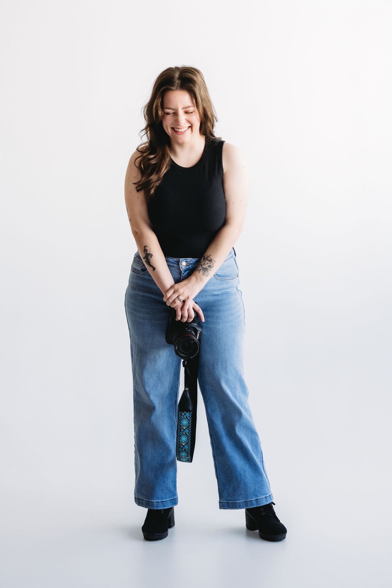 Person wearing denim jacket and black top sits cross-legged on white background while holding camera.