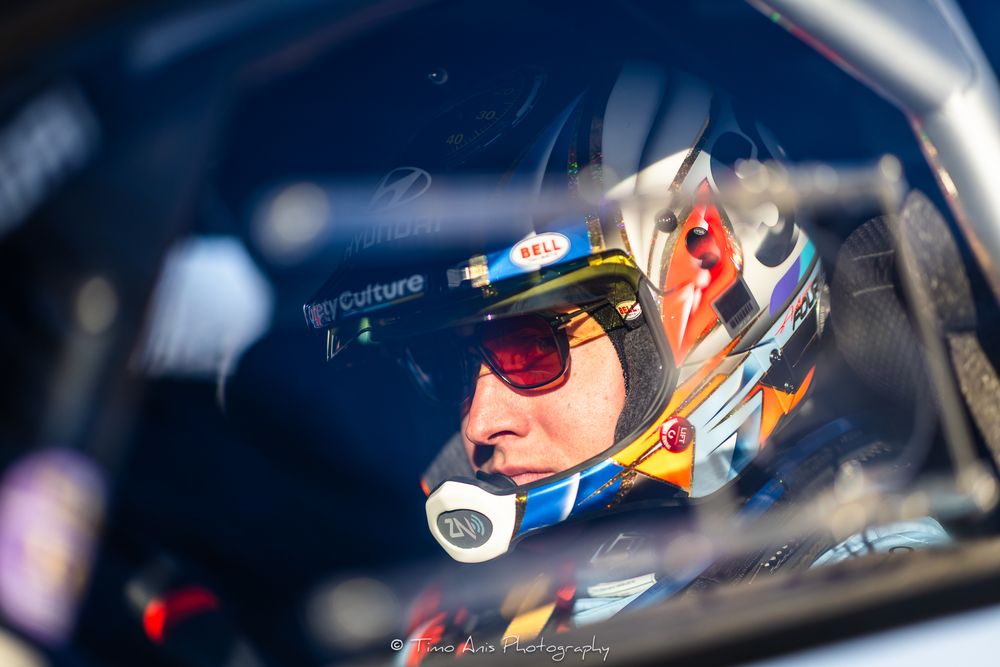 Race car driver wearing sunglasses and a racing helmet sits in cockpit of vehicle during competition.