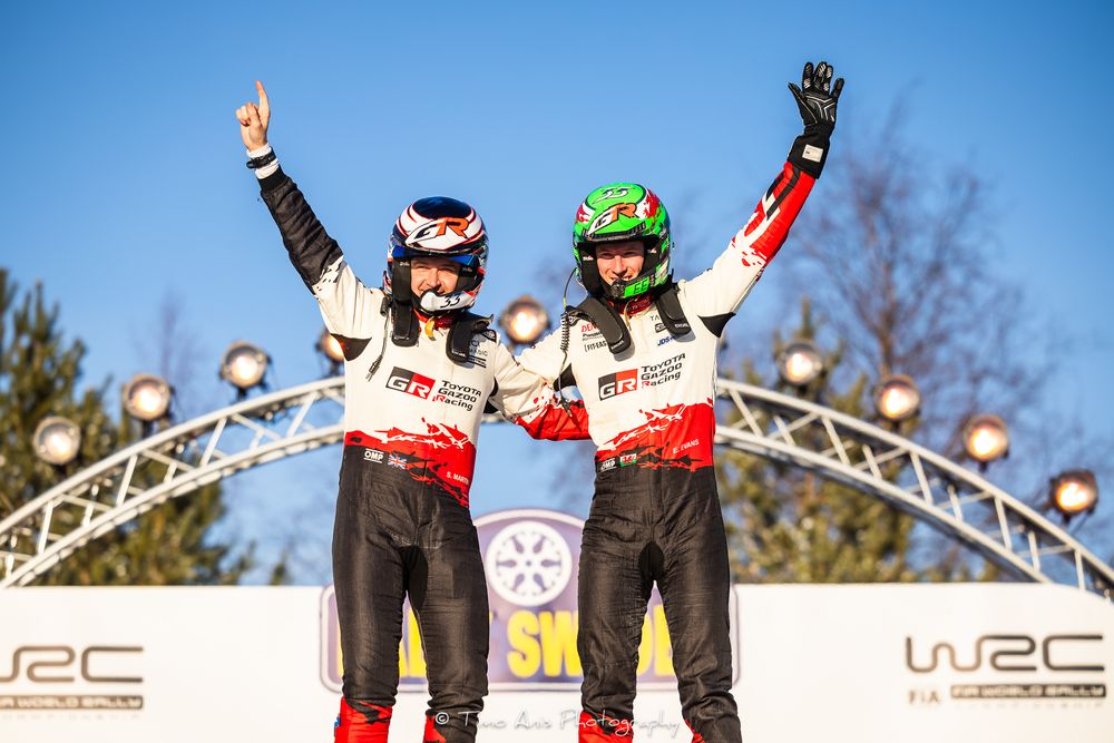 Rally drivers celebrate on podium platform after victory with champagne spray and trophies at WRC championship event.