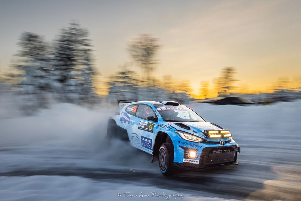 Rally car racing through snowy terrain at sunset with motion blur and dramatic lighting effects.
