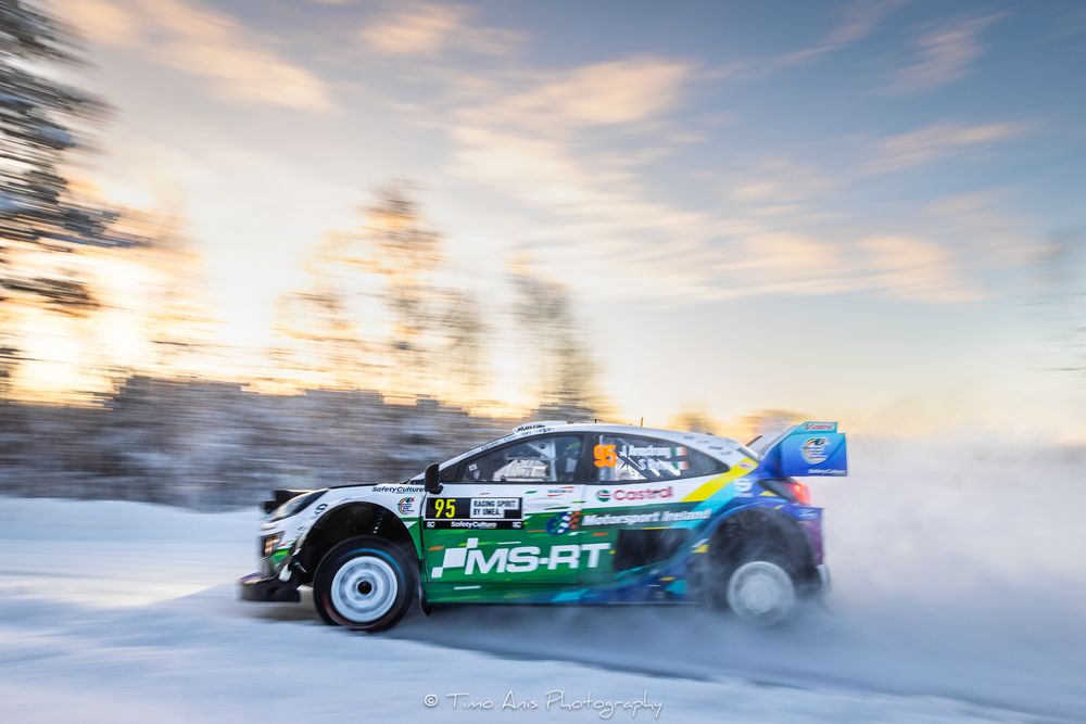 Rally car racing through snowy terrain at sunset with motion blur and dramatic sky in winter conditions.