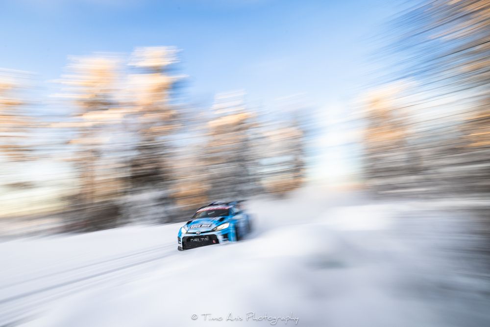 Blue rally car drifts on snow-covered course through forest with artistic motion blur effect.