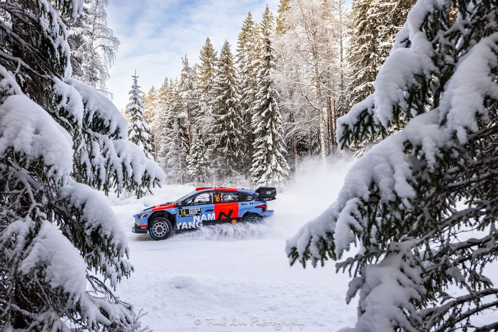 A snowy forest road curves through snow-covered evergreen trees with a blue car visible in the distance.