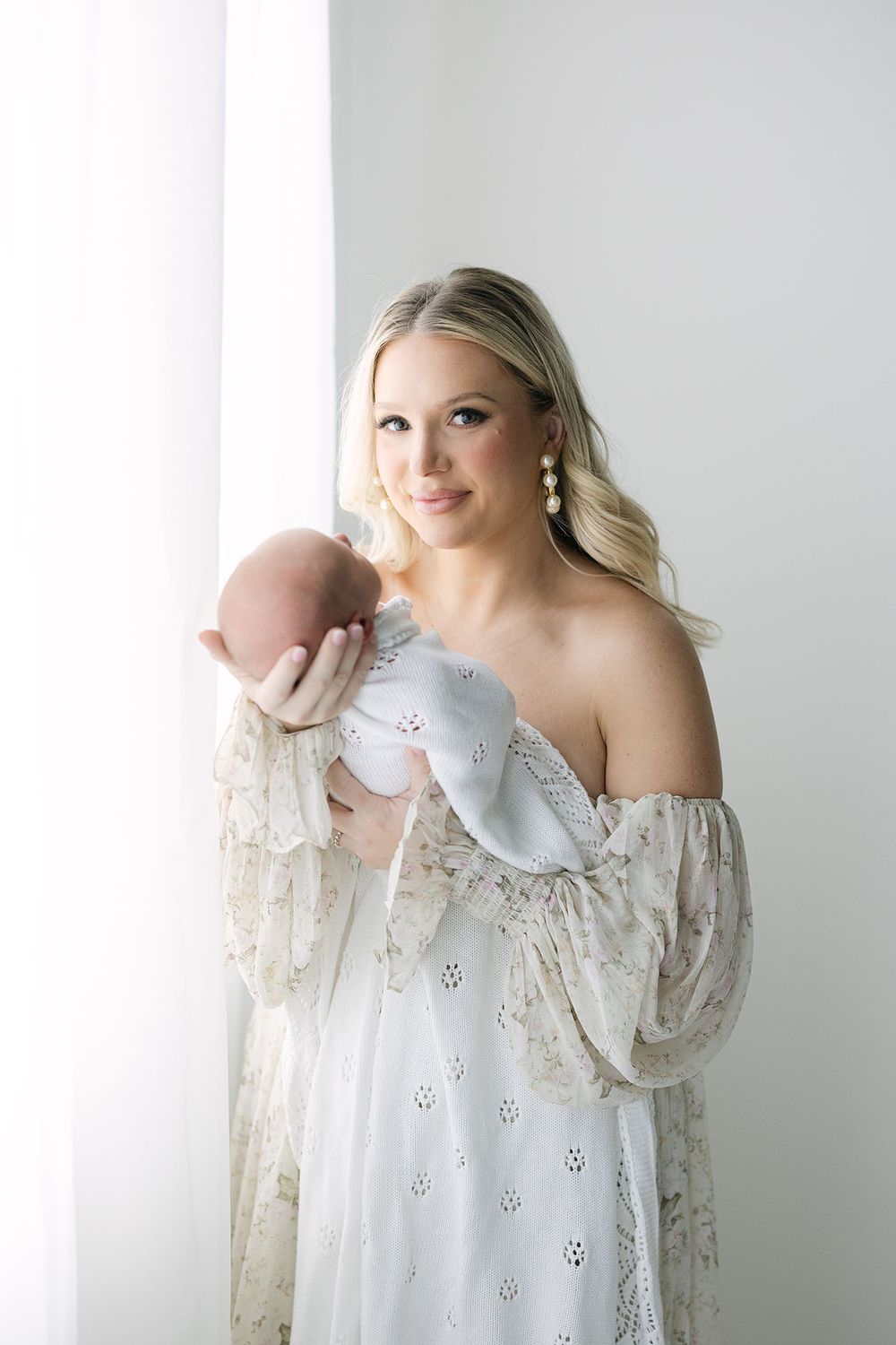 Mom standing next to a window while holding her baby in an all white studio by Alex Ishee Photography.