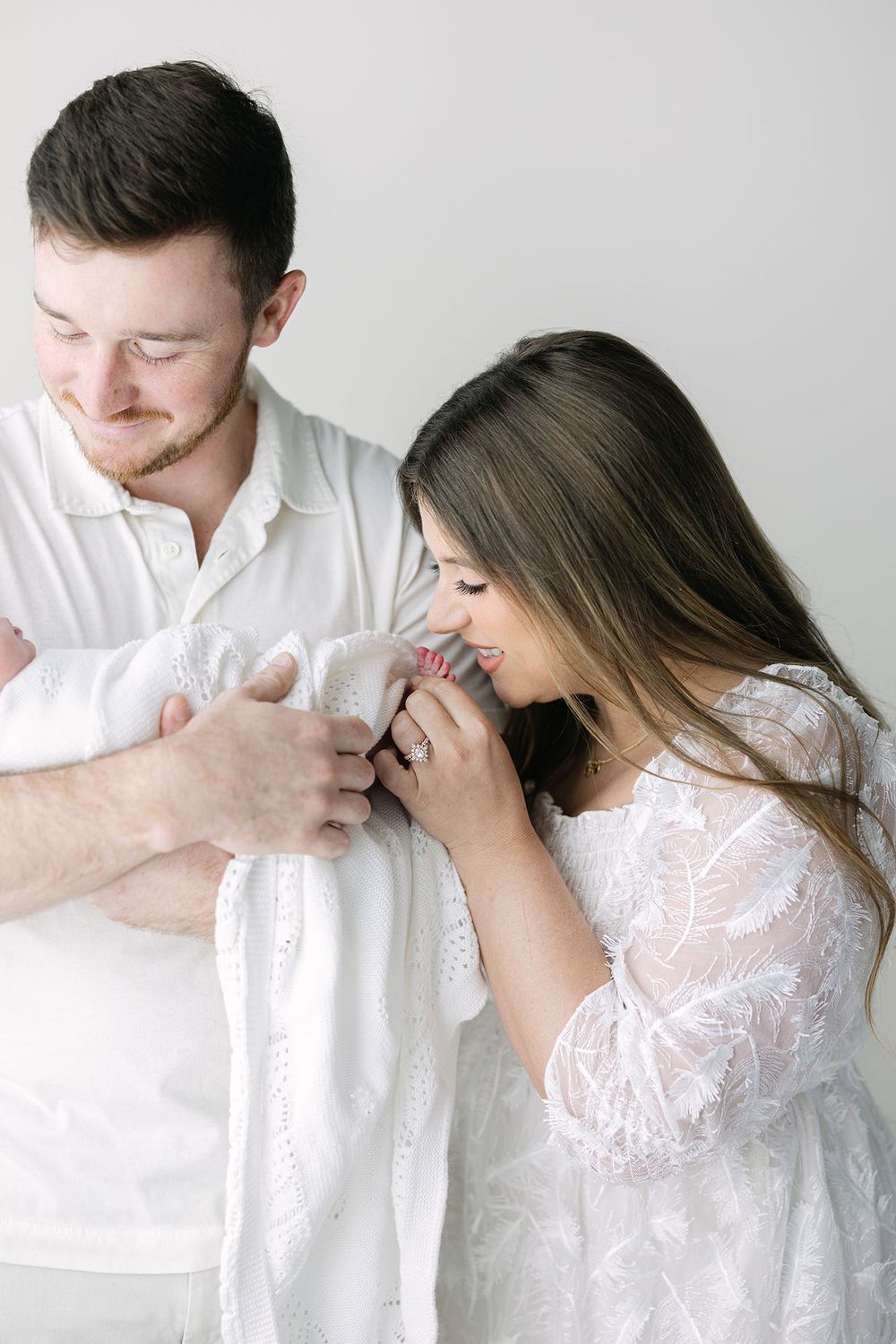 Dad holding a baby in white swaddles while mom kisses baby's toes in a white studio.