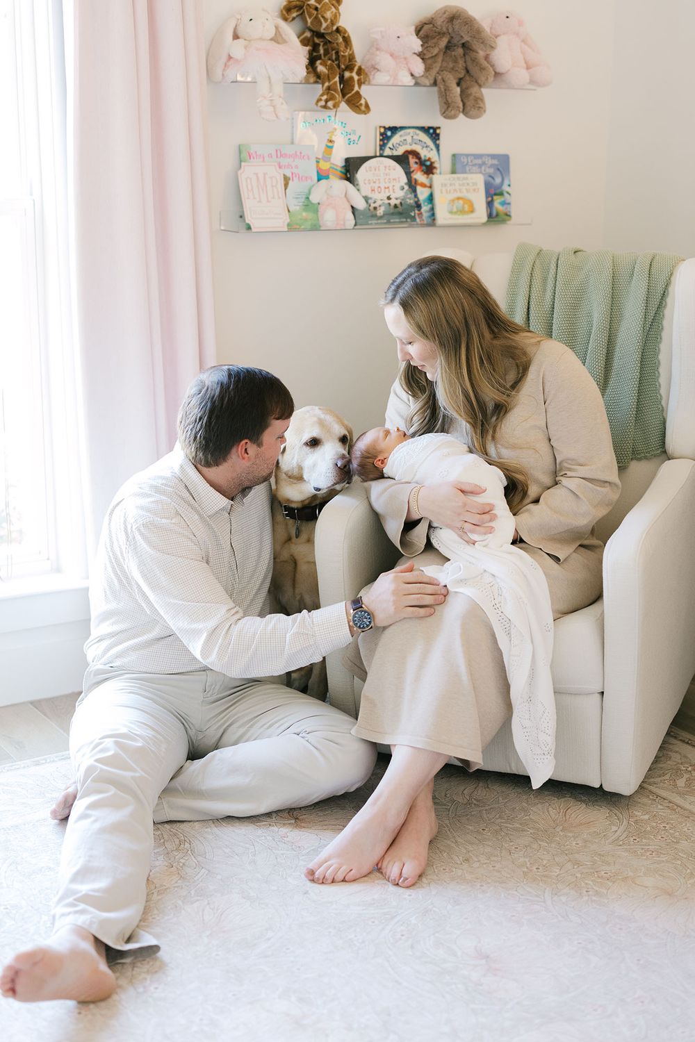 Mom holding baby in a nursery rocking chair while dad sits on the floor and kisses a yellow lab.