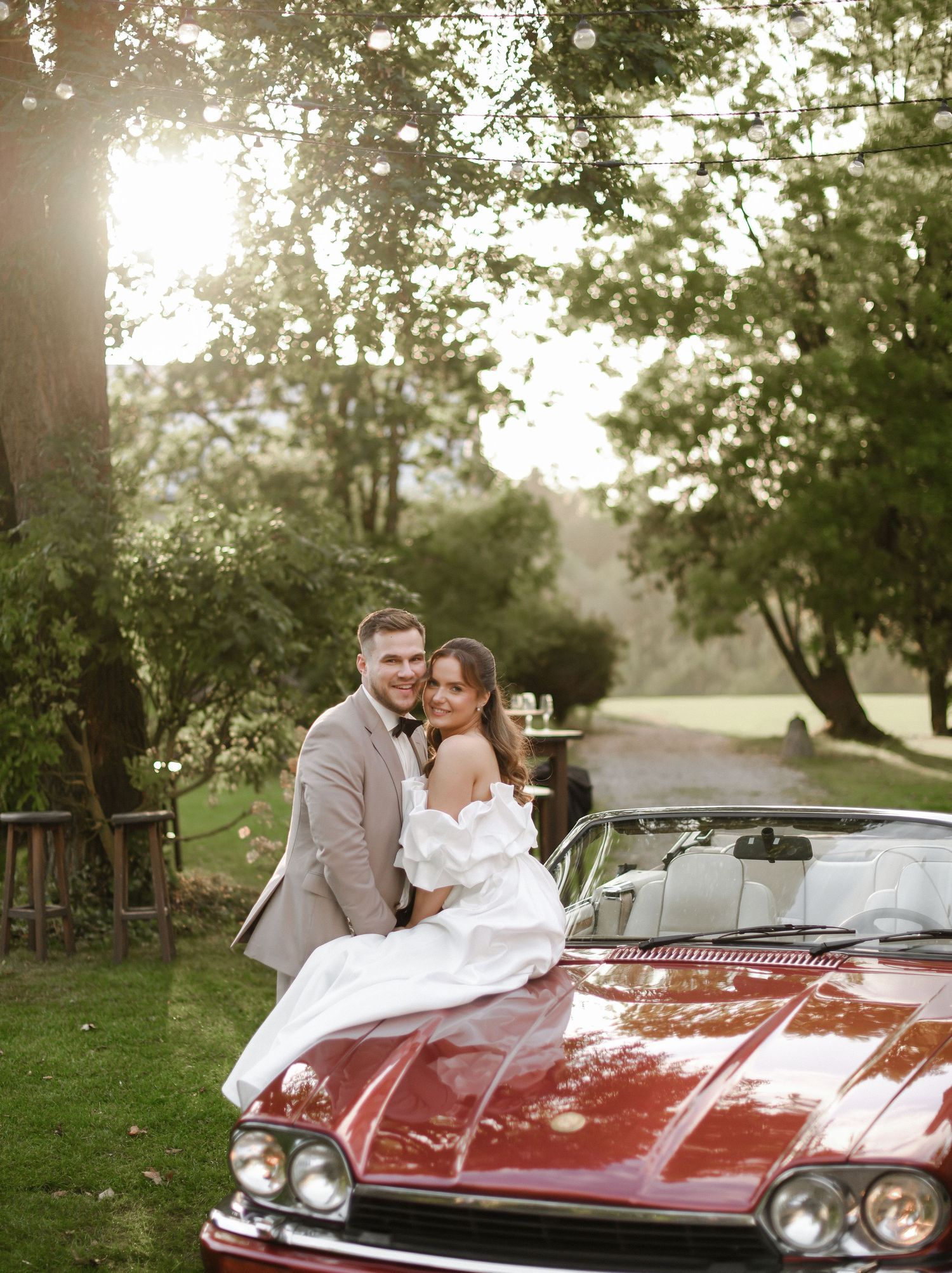 A couple sits together in a classic red vintage Cadillac convertible on a sunny afternoon in a park setting.
