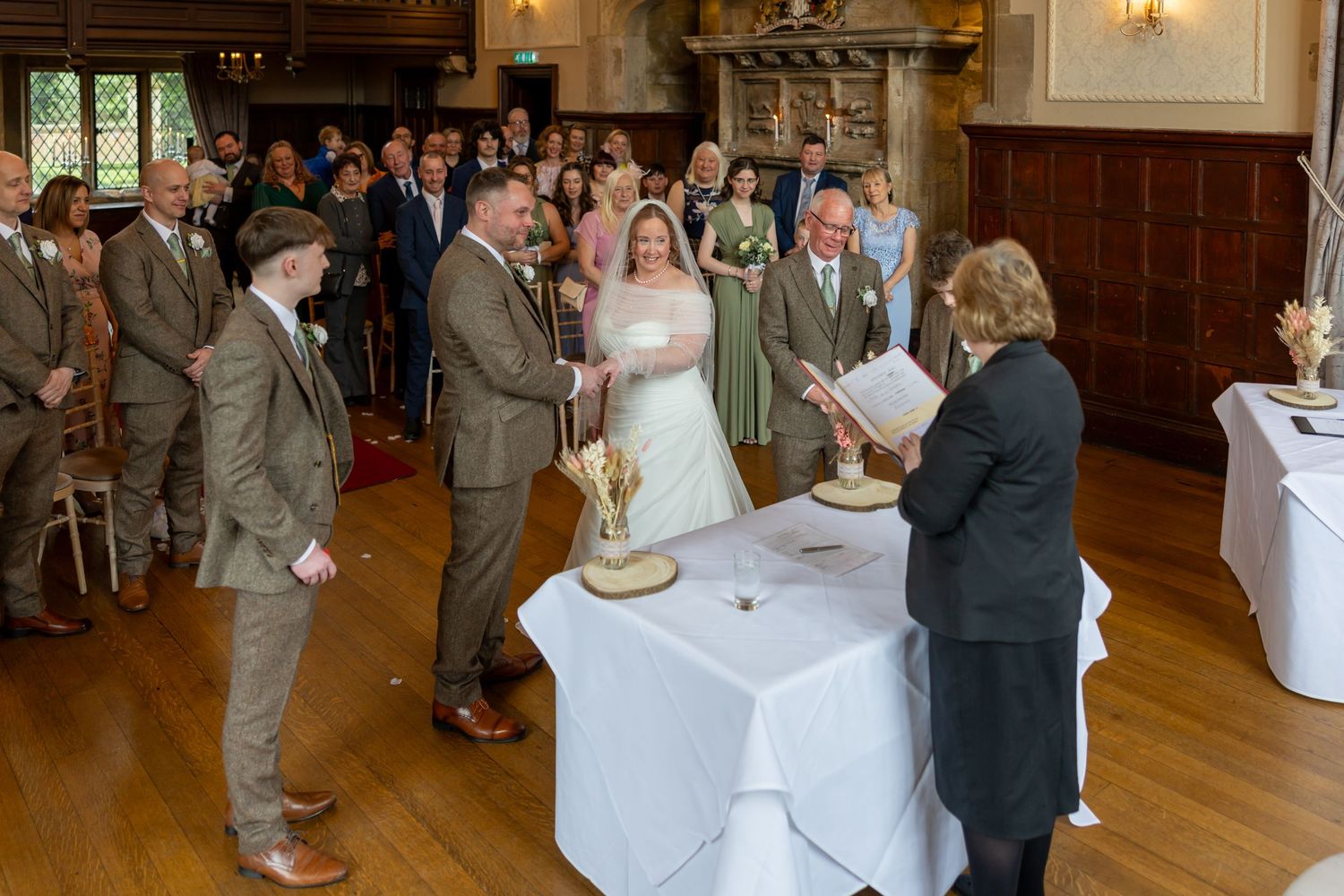 Newlyweds at a wedding ceremony sign documents with officiant at wooden-paneled venue with guests watching.