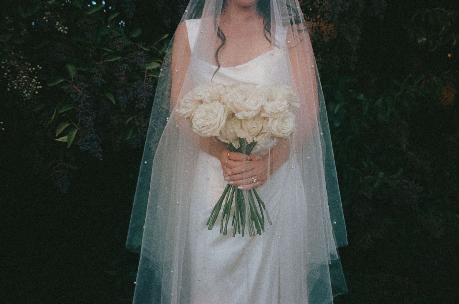 A bride in a white wedding dress holds a large bouquet of cream-colored roses against a dark green garden background.