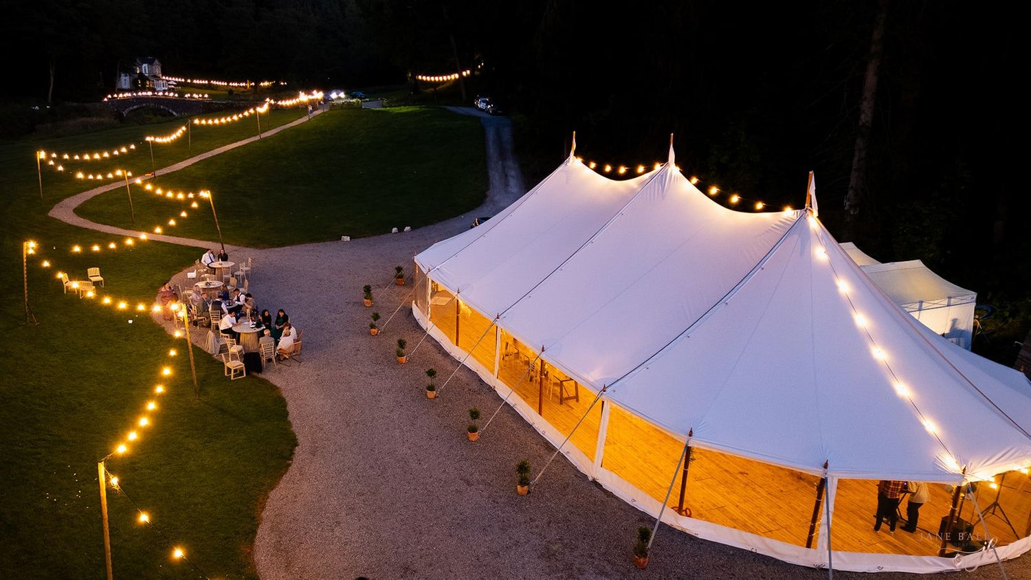 A white wedding tent illuminated with string lights glows against the evening sky during an outdoor reception.