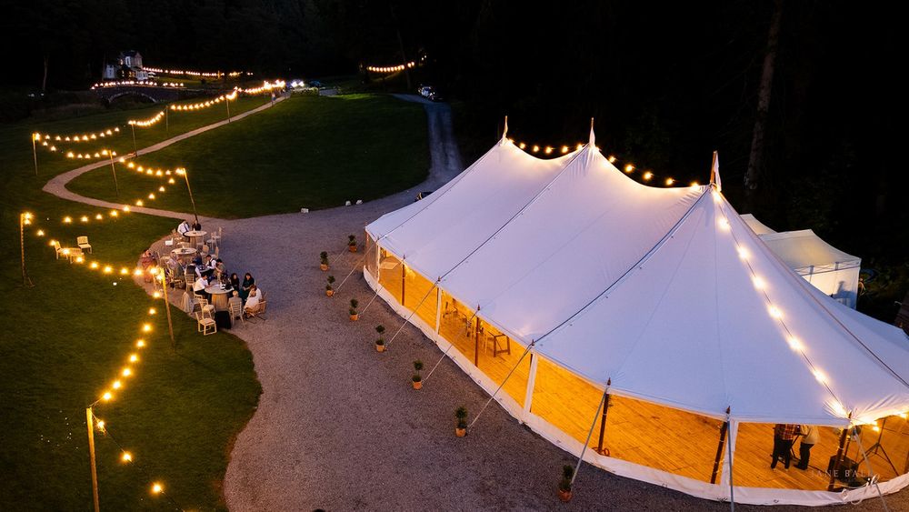 A white wedding tent illuminated with string lights glows against the evening sky during an outdoor reception.