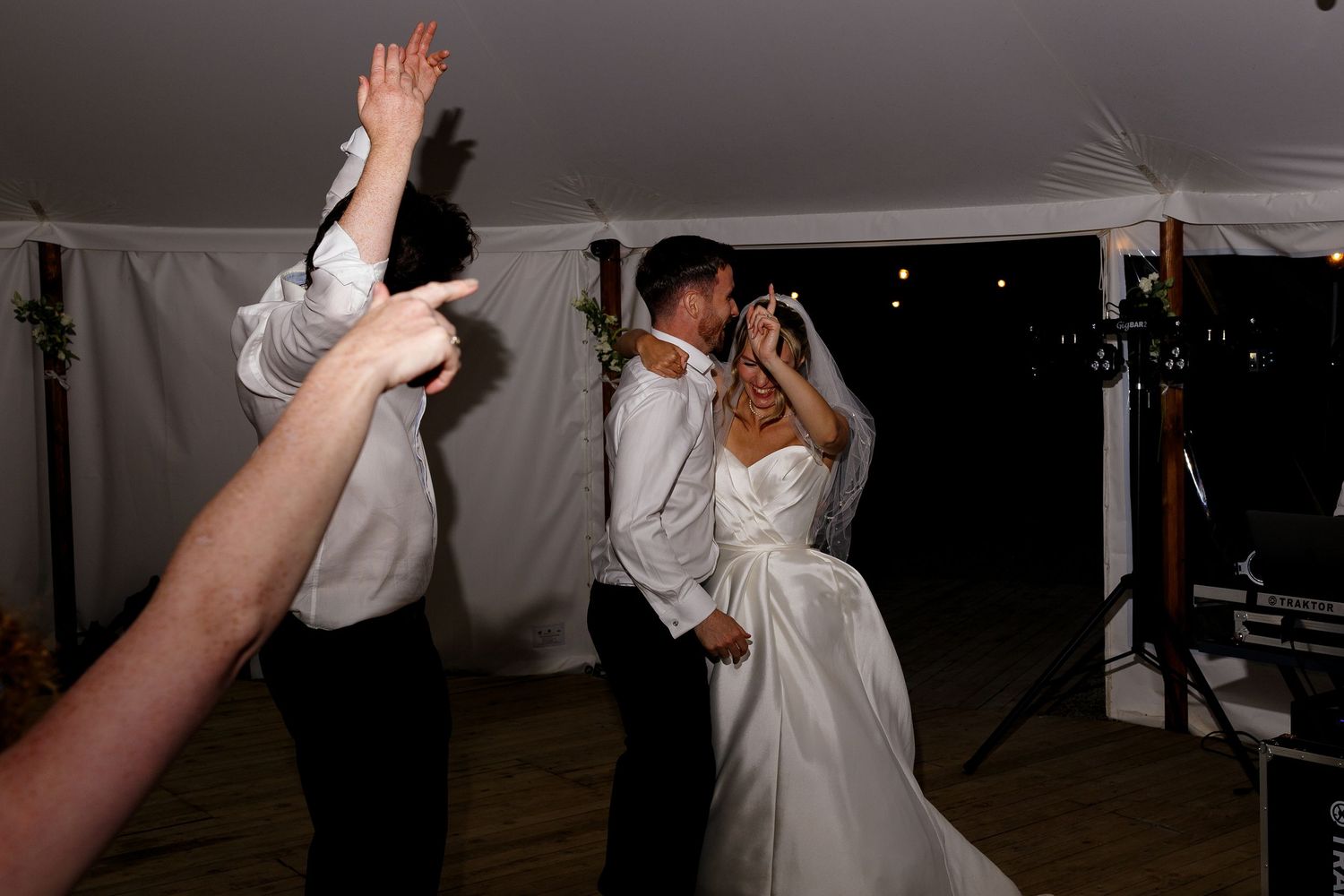 Wedding guests dancing and celebrating at a reception under a tent with arms raised in joyful movement.