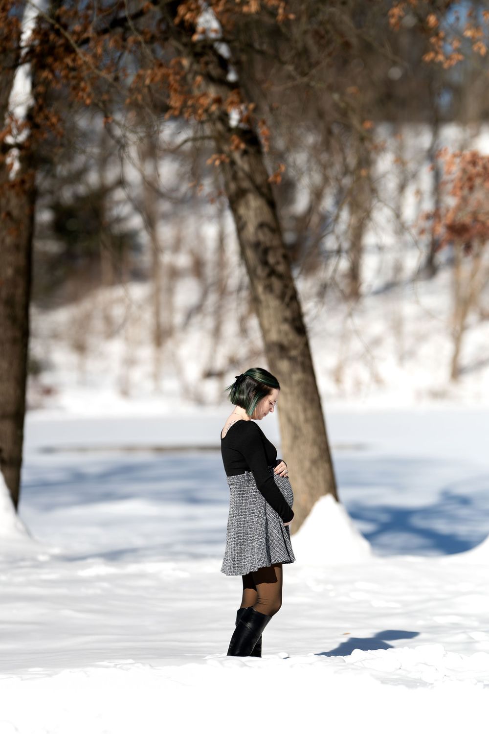 Pregnant woman in a plaid skirt and black top standing sideways in a snowy winter park setting.