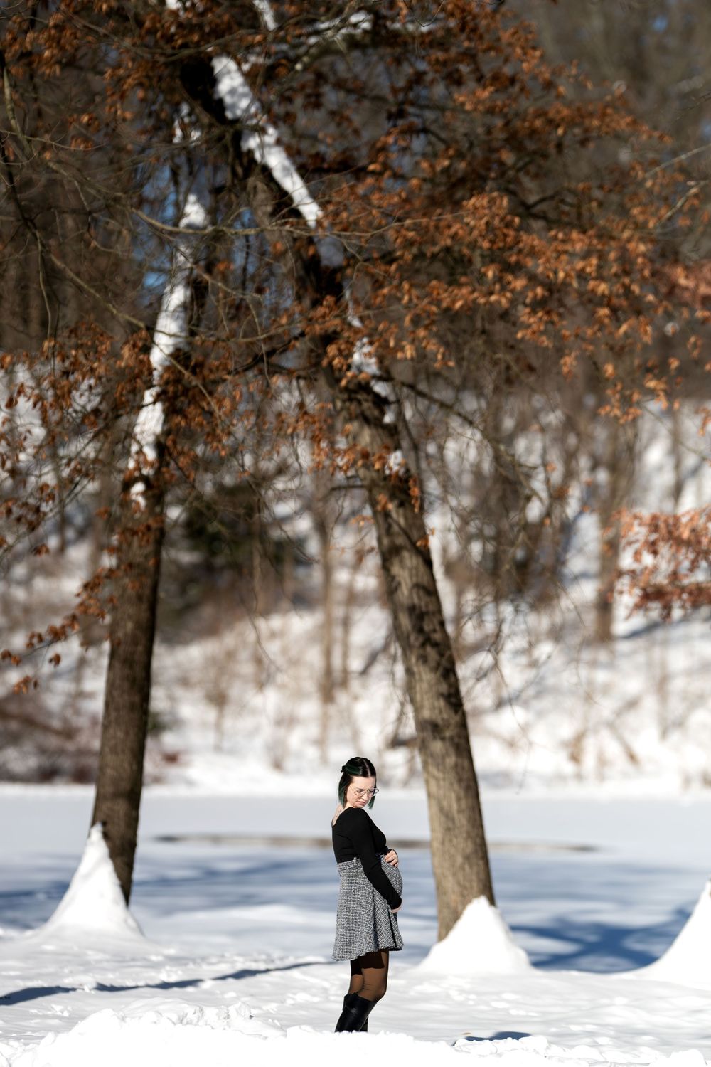 Pregnant woman in plaid skirt posing beside a birch tree in a bright snowy winter park.
