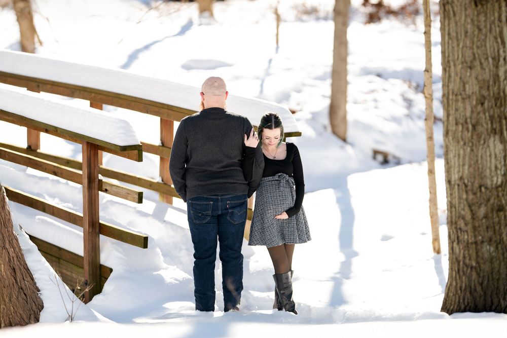 Couple walking away together through snowy winter landscape beside wooden bridge, woman in plaid skirt and knee-high boots.