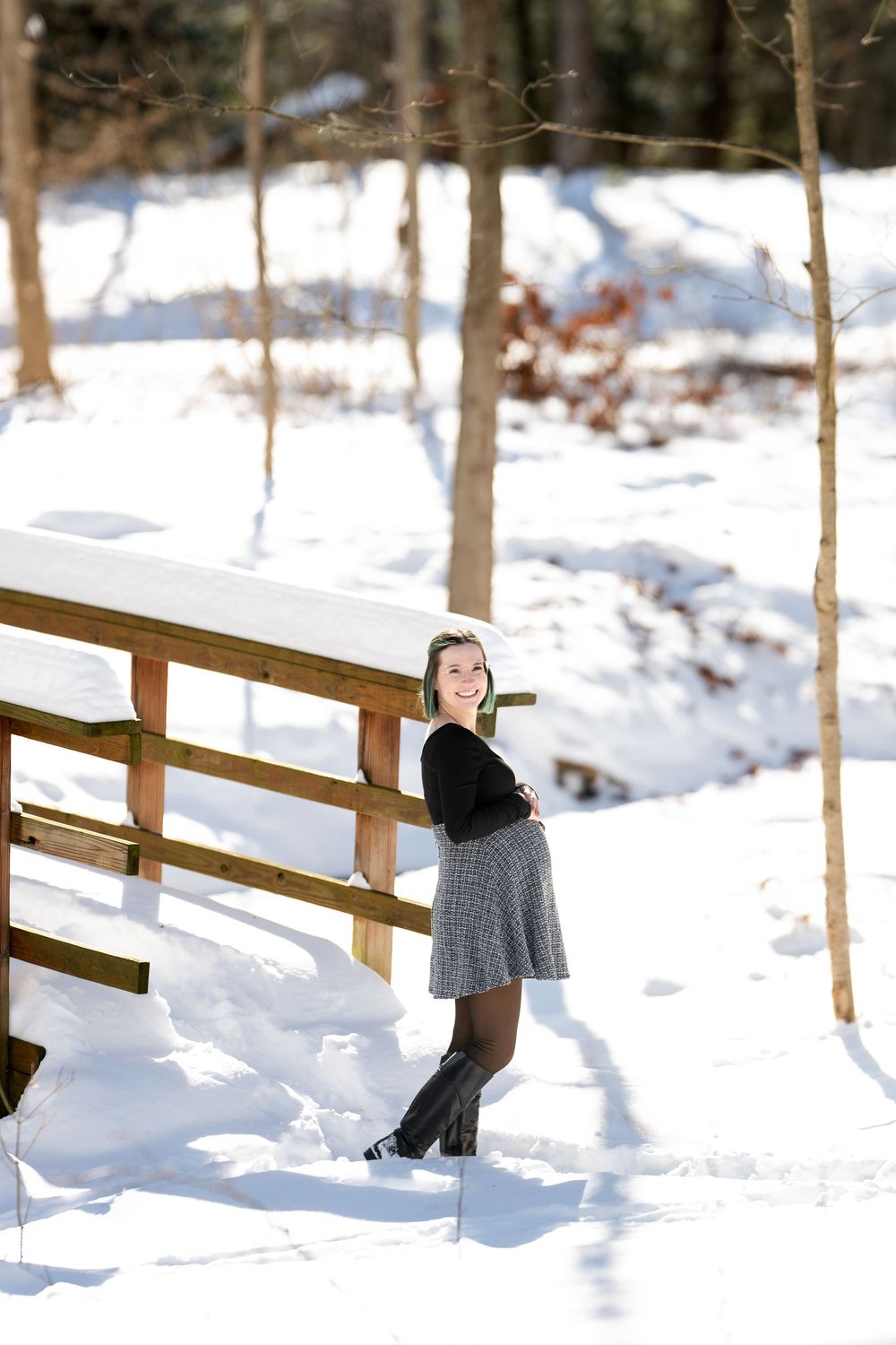 Smiling pregnant woman in black top and plaid skirt posing beside snow-covered wooden bridge in sunny winter forest.