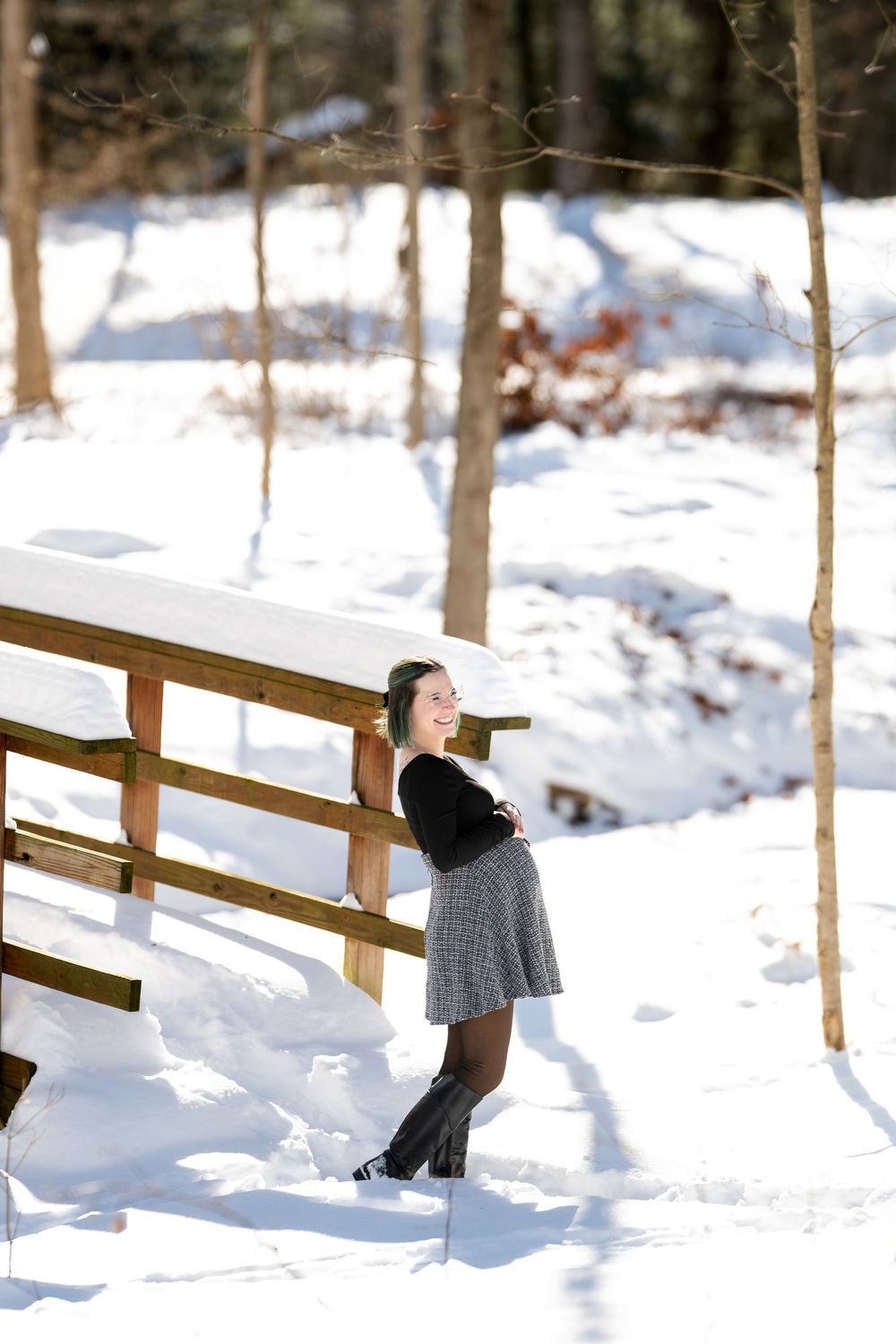Pregnant woman in plaid skirt and black top leaning against snow-covered wooden fence in bright winter forest setting.