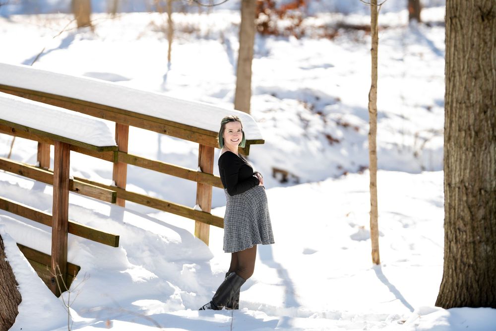 Pregnant woman in black long-sleeve top and plaid skirt standing beside rustic wooden bridge in snow.