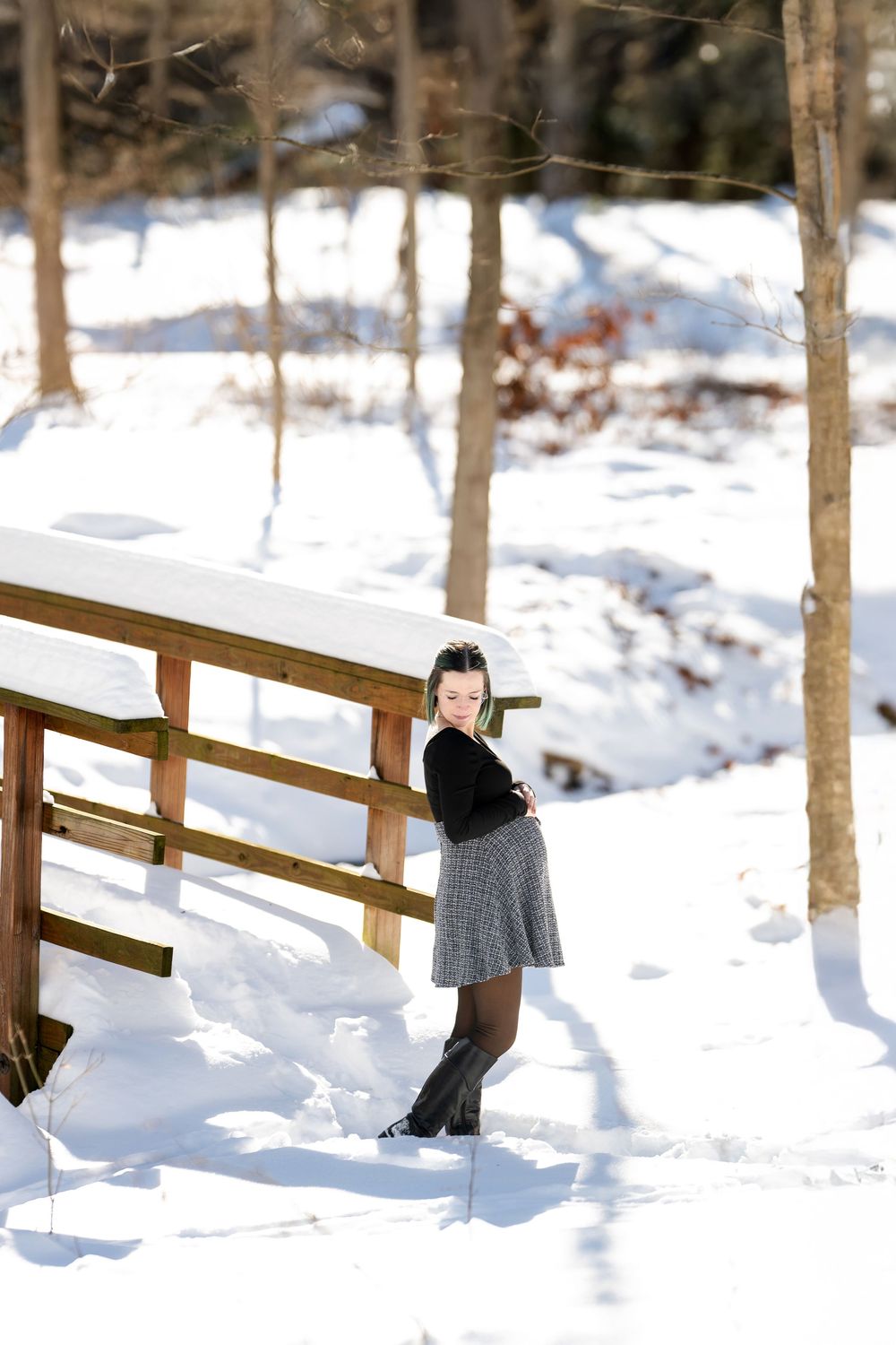 Woman in plaid skirt and black top posing beside snow-covered wooden bridge, looking back over shoulder in winter forest.