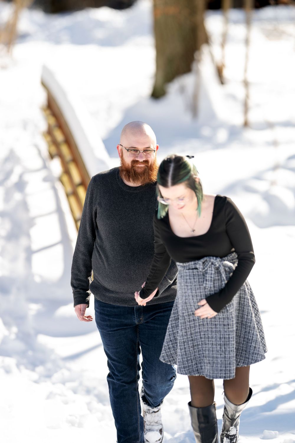 Laughing couple walking through snowy landscape near wooden bridge, man with red beard and woman with green-tinted hair.