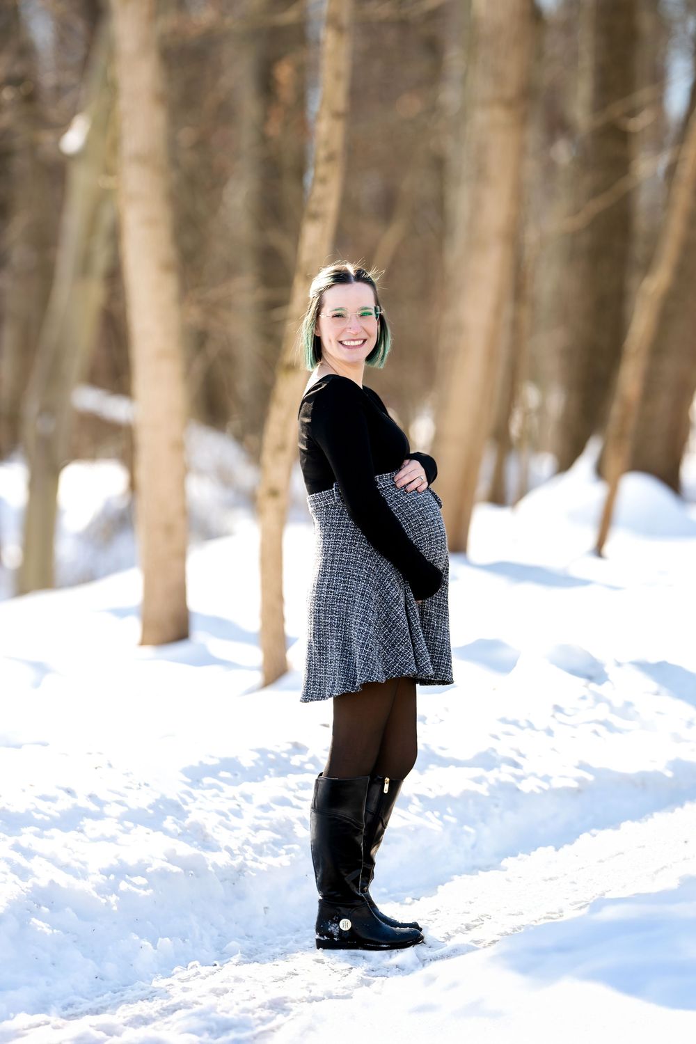 Happy pregnant woman in black top, plaid skirt, and tall black boots posing in sunny snowy forest.