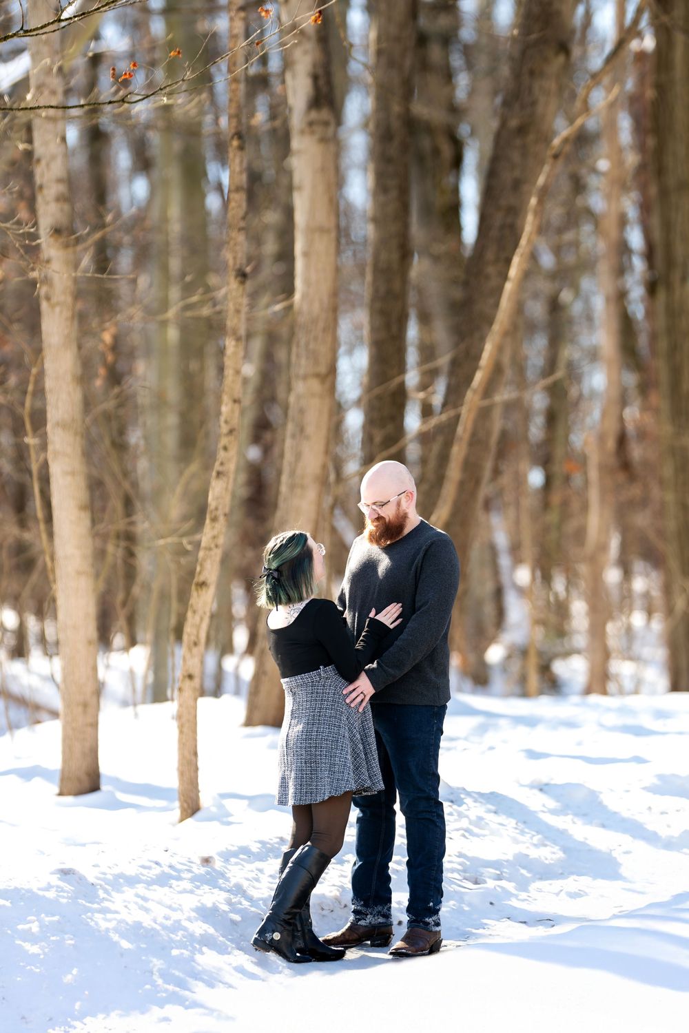 Couple standing together in snowy winter forest, woman with green hair in plaid skirt facing bearded man.