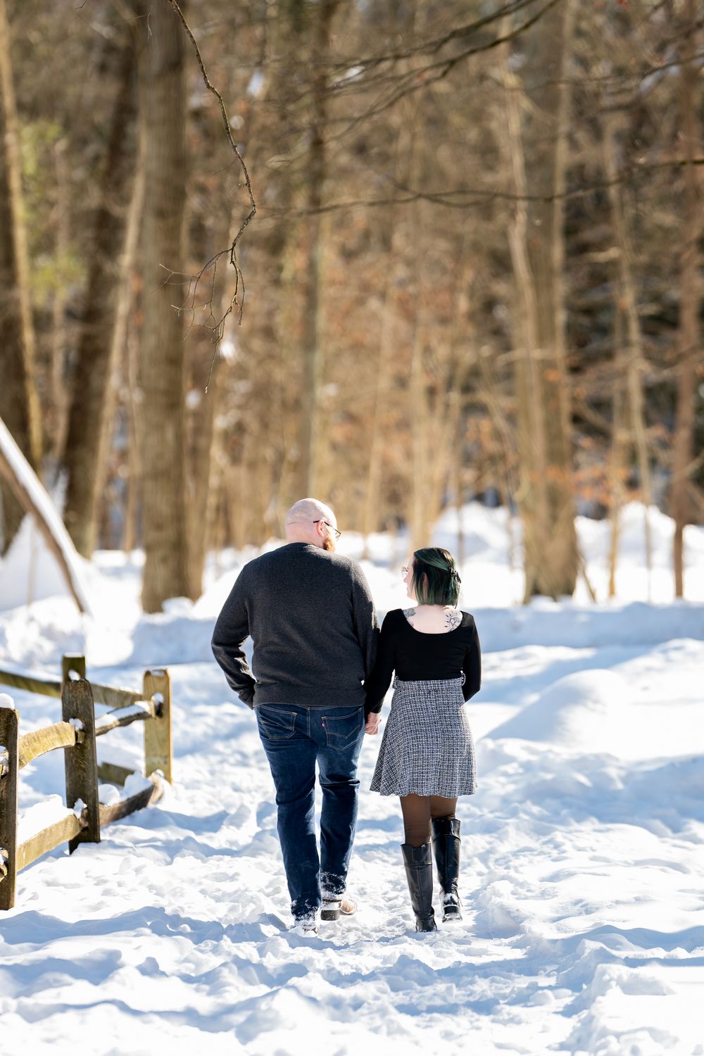 Couple walking away hand in hand down snowy forest path beside rustic wooden fence in winter.