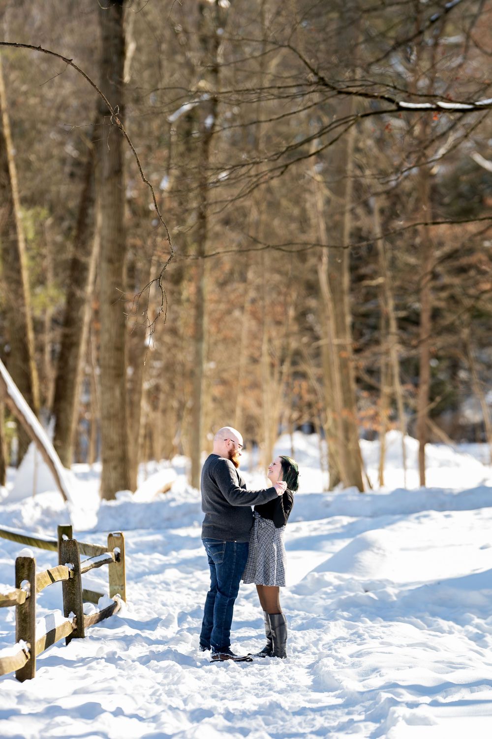 Couple holding hands and dancing on a snowy forest path next to a wooden fence on a sunny winter day.