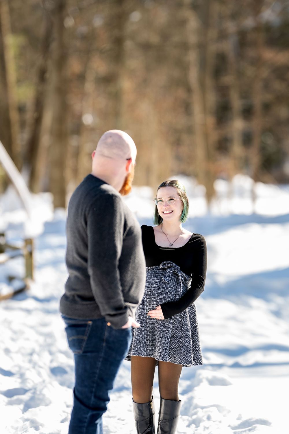 Smiling pregnant woman in a plaid skirt and knee-high boots facing her partner on a snowy forest path.
