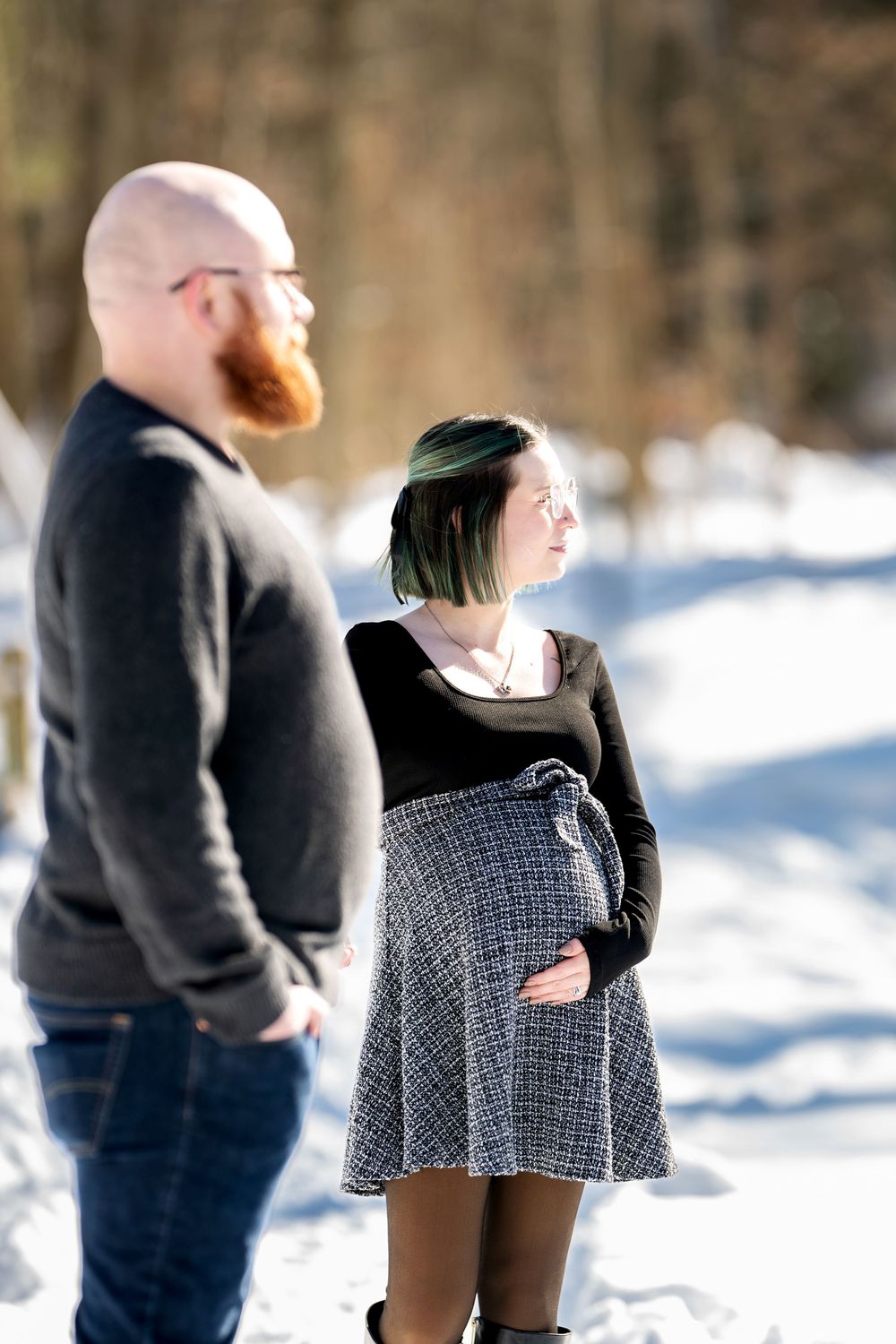 Pregnant woman in a black top and plaid skirt cradling her baby bump beside her bearded partner in snow.