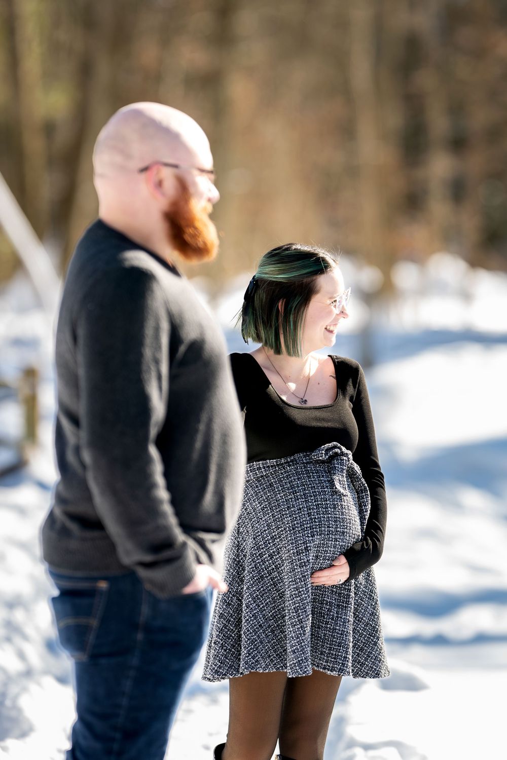 Laughing pregnant woman with green-tipped hair holding her baby bump while standing with her partner in snow.