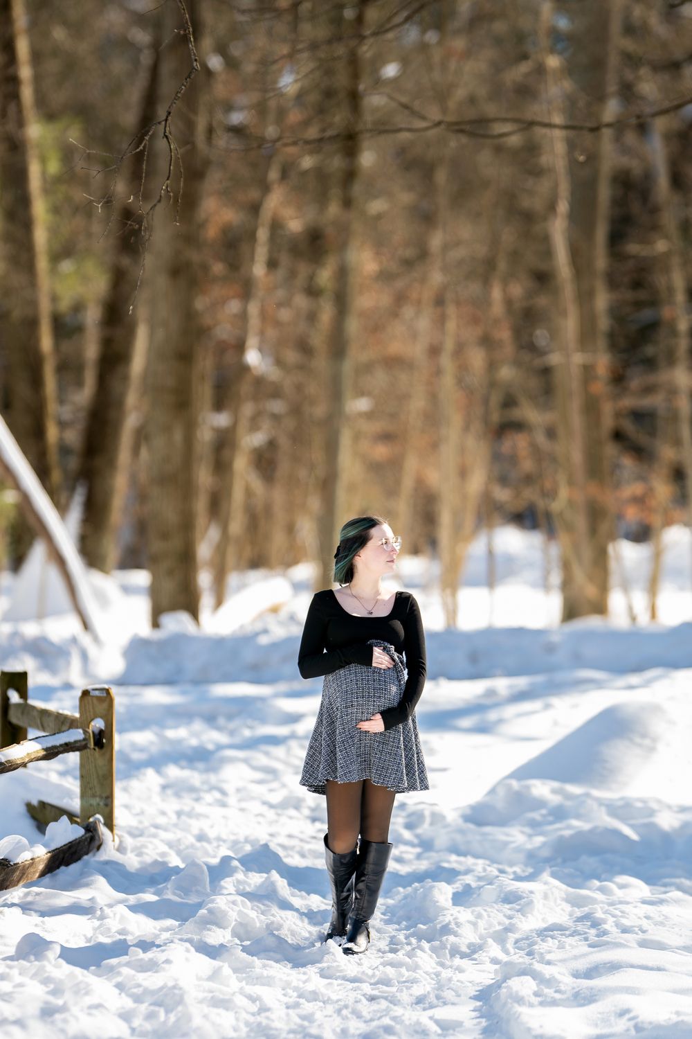 Pregnant woman with green-highlighted hair laughing while standing on a sunny snow-covered forest path.