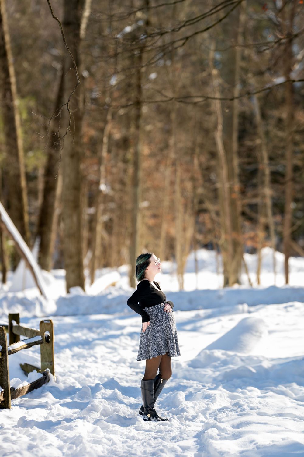 Woman in skirt and knee-high boots standing in snowy winter forest looking up at the sky.