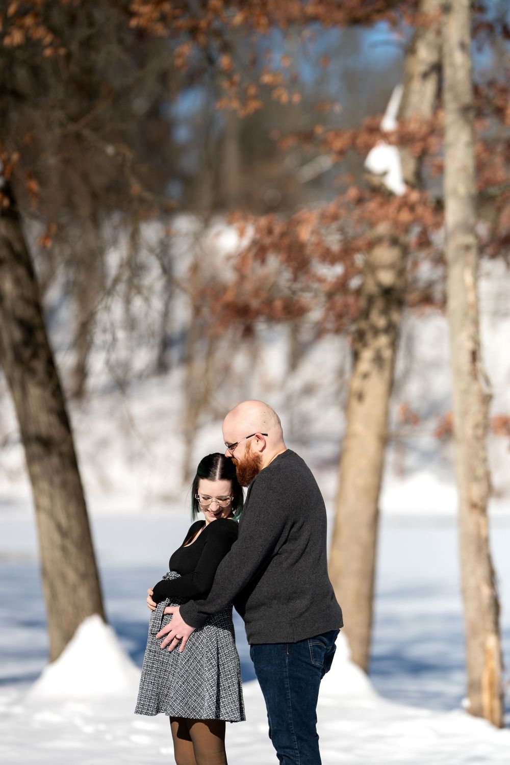 Bearded man tenderly touching pregnant partner's belly while she smiles in a snowy winter forest.